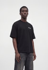 Junger Mann trägt ein schwarzes oversized T-Shirt mit dem Schriftzug „Dropsiz“ und ausgewaschene schwarze Jeans, steht vor einem schlichten weißen Hintergrund.