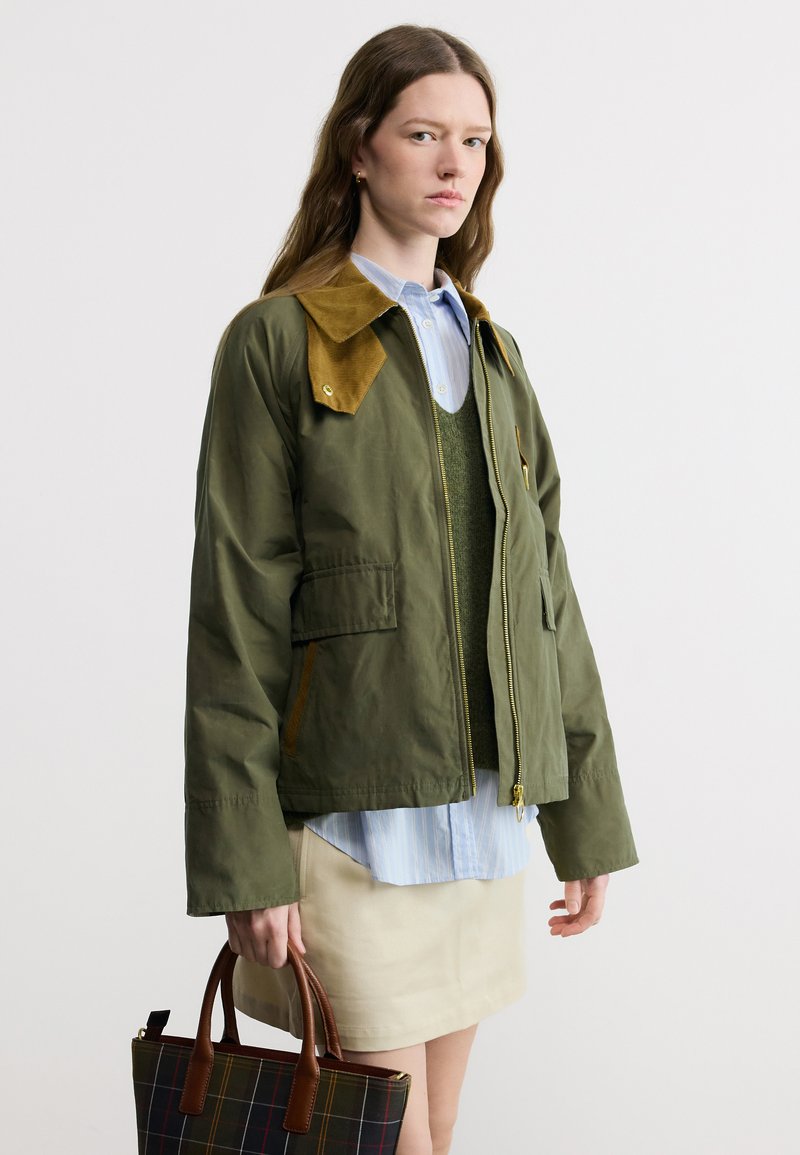 Young woman in olive green jacket over blue shirt and beige skirt holding a plaid handbag against white background.