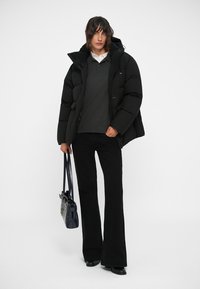 Casaco puffer preto com capuz, sobre um suéter cinza escuro e uma camisa branca com colarinho. Usado com calças pretas de boca larga e a segurar uma mala escura.