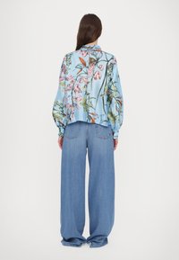 Donna che indossa una camicia a maniche lunghe con stampa floreale blu e jeans larghi blu, in piedi davanti a uno sfondo bianco semplice.