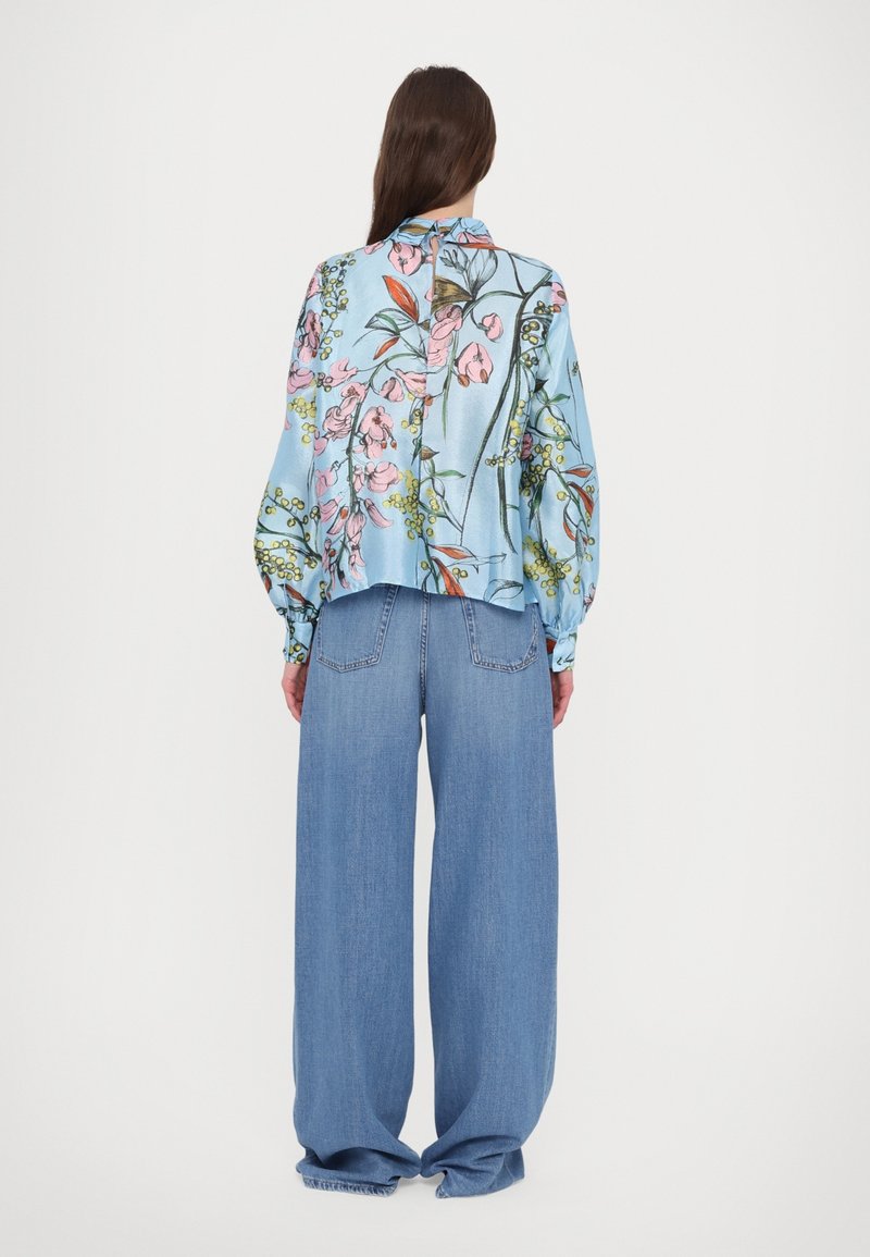 Donna che indossa una camicia a maniche lunghe con stampa floreale blu e jeans larghi blu, in piedi davanti a uno sfondo bianco semplice.