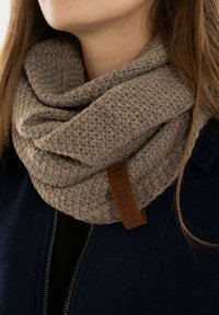 Gehaakte taupe sjaal met een gestructureerde weving, voorzien van een bruine leren label. Gedragen over een donkere jas, waarbij het gewikkelde ontwerp en de dikte goed tot uiting komen.