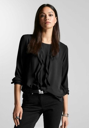 Schwarze Bluse mit Rüschendetail an der Vorderseite, lange Ärmel, die an den Manschetten hochgerollt sind, kombiniert mit schwarzen Jeans und einem silbernen Ring. Einfaches, elegantes Design.