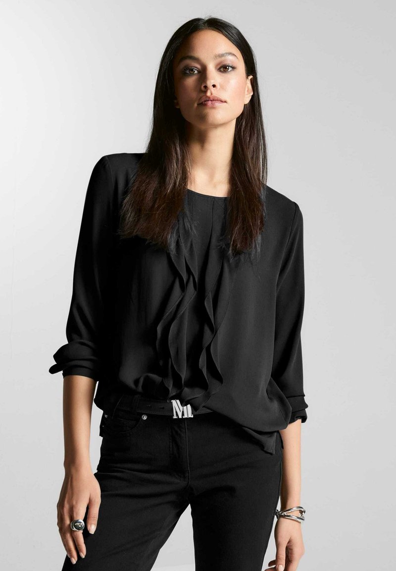 Zwarte blouse met gerimpelde voorkant, lange mouwen opgerold bij de manchetten, gecombineerd met zwarte jeans en een zilveren ring. Eenvoudig, elegant ontwerp.