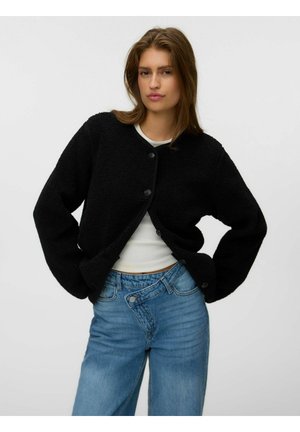 Ung kvinde iført en sort fleecetrøje med knapper, hvid top og højtaljede blå jeans, stående med hænderne i siden.