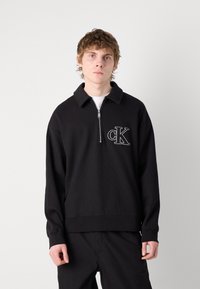 Black zip-up sweatshirt with a ribbed texture and a collar. Features a white embroidered "cK" logo on the left chest.