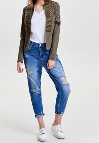 Chaqueta militar de color verde oliva con botones decorativos, combinada con jeans azules desgastados. Una camiseta blanca y zapatillas blancas completan el conjunto.