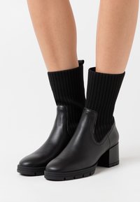 Mustat nahkasaappaat nilkoille, joissa on ribatoidut mustat sukkamaiset jatkeet, lohkareet, pyöristetyt varpaat ja tekstuuripohjat pitoa varten.