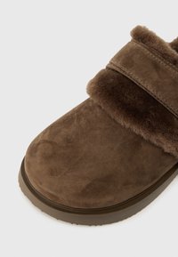 Pantofii slip-on din suede maro, cu talpă groasă din cauciuc și căptușeală din blană pufoasă în jurul deschiderii, concepuți pentru căldură și confort.