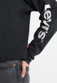 Czarny długi rękaw bluzka z kolekcji „Levi's” w srebrnym kolorze na rękawie. Noszona z szarymi dżinsami z widocznym detalem miedzianego guzika.