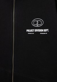 Schwarzes Kleidungsstück mit Frontreißverschluss, weißer Text "PROJECT DIVISION DEPT." unter einem runden Logo mit den Initialen "PD" und "1991" darin.