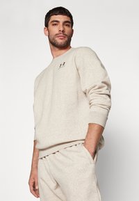 Sudadera beige de tela suave con cuello redondo y puños acanalados, que presenta un pequeño logo negro en el pecho. Combinada con pantalones a juego.