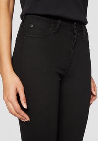 Primer plano de una persona que lleva unos pantalones vaqueros negros de cintura alta con bolsillos frontales visibles y una cremallera parcialmente subida.