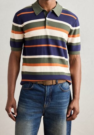 Man draagt een poloshirt met korte mouwen in marineblauw, olijfgroen, oranje en wit gestreept, een blauwe spijkerbroek en een beige riem, staand tegen een effen achtergrond.