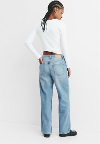 Široke jeans hlače v modri denimu z zadnjimi žepi, ki vsebujejo svetlo rjavo usnjeno nalepko. Usklajene z oprijetim belim dolgim rokavom.