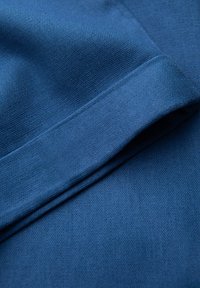 Tissu bleu avec un tissage texturé, présentant un bord replié et une finition lisse. Des coutures visibles le long du pli ajoutent du détail.