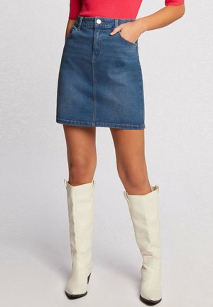 Jupe en denim bleu moyen, design ajusté avec une fermeture à bouton sur le devant, deux poches avant et un ourlet droit. Associée à des bottes blanches montantes.