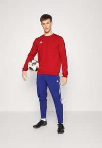 Sweat-shirt rouge Adidas à manches longues, associé à un jogging bleu Adidas avec des rayures blanches. Un ballon de football noir sous un bras. Des crampons noirs.