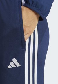 Marineblauwe sportbroek met een wit drie-strepen ontwerp aan de zijkant, met een hand die rust in een zijzak voor detail.