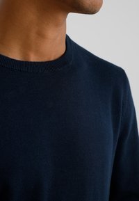 Zbliżenie osoby noszącej ciemnogranatowy sweter z okrągłym dekoltem na jasnoszarym tle.