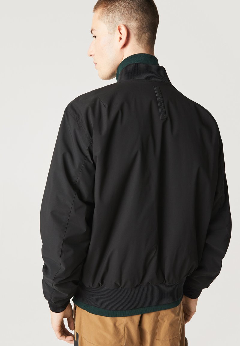 Homme portant une veste bomber noire avec un col, des poignets et un ourlet côtelés, associé à un pantalon beige, tourné le dos contre un fond uni.