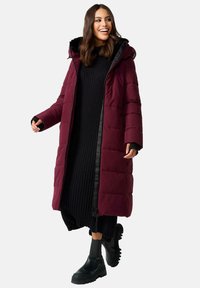 Burgundijas pūkains mētelis ar kapuci, ar rāvējslēdzēju priekšpusē, ar quilted tekstūru un melnām akcentiem, apvienots ar melnu ribotu kleitu un zābakiem.
