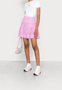 Gonna eyelet rosa con orlo arricciato, abbinata a una maglia bianca a maniche corte. Gli accessori includono una piccola borsa rosa chiaro e sneaker bianche.