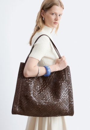 Donna con capelli chiari che indossa un top bianco a coste e tiene una grande borsa in pelle marrone intrecciata sulla spalla.