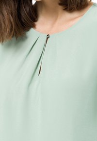 Blusa verde chiaro con scollatura rotonda, caratterizzata da un bottone decorativo nella parte anteriore e sottili pieghe per aggiungere texture. Tessuto morbido.