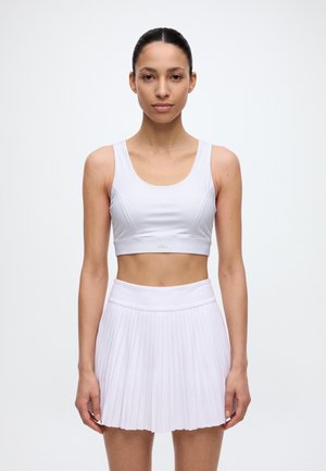 Une femme présente un soutien-gorge de sport blanc et une jupe de tennis plissée sur un fond clair et uni.