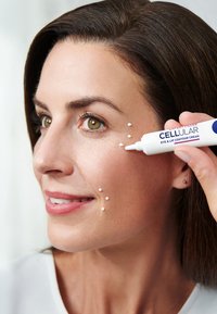 White eye and lip contour cream being applied to the cheek, with small dots visible. The product tube has blue and red text on a white background.