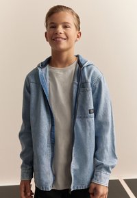 Veste en jean bleu clair avec capuche, dotée d'une fermeture éclair et d'une poche poitrine. Portée sur un t-shirt gris clair. Design décontracté.