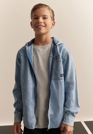 Hellblaue Jeansjacke mit Kapuze, ausgestattet mit einem Reißverschluss und einer Brusttasche. Darüber getragen wird ein hellgraues T-Shirt. Lässiges Design.