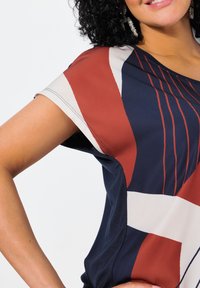 Blusa de manga corta con un patrón geométrico en azul marino, rojo y crema. Tejido suave con un corte relajado y detalle de costura sutil.