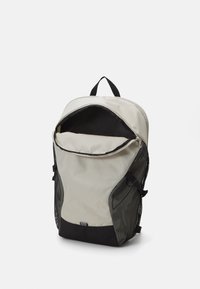 Sac à dos avec un corps en tissu gris clair, des poches latérales en maille noire et une fermeture à glissière. Dispose d'une poignée supérieure pour le transport.