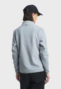 Svijetloplava flis jakna s mekim teksturom, s uzdignutim ovratnikom i crnom kapom sa logotipom. Stražnji pogled prikazuje opušteni kroj.