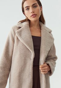 Abrigo oversized de pelo sintético color beige con cuello de muesca, superficie texturizada y bolsillos laterales, llevado sobre una parte de arriba oscura.