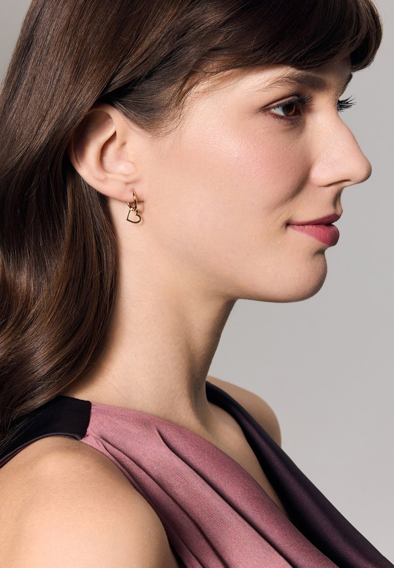 Złote kolczyki w kształcie kół z zawieszką w kształcie serca, z polerowanym wykończeniem. Modelka ma gładkie, średnio brązowe włosy i nosi fioletową odzież.