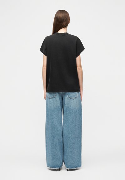 Γυναίκα που στέκεται με την πλάτη προς τα εμάς, φοράει μαύρο τοπ με κοντά μανίκια και ανοιχτό μπλε τζιν παντελόνι με φαρδιές μπατζάκια και ξεφτισμένα τελειώματα.
