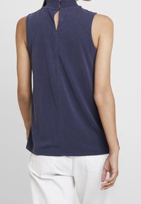 Top sin mangas de color navy con cuello alto, que presenta una abertura tipo ojal en la parte posterior. Tela suave, corte relajado, con detalles de costura limpias.