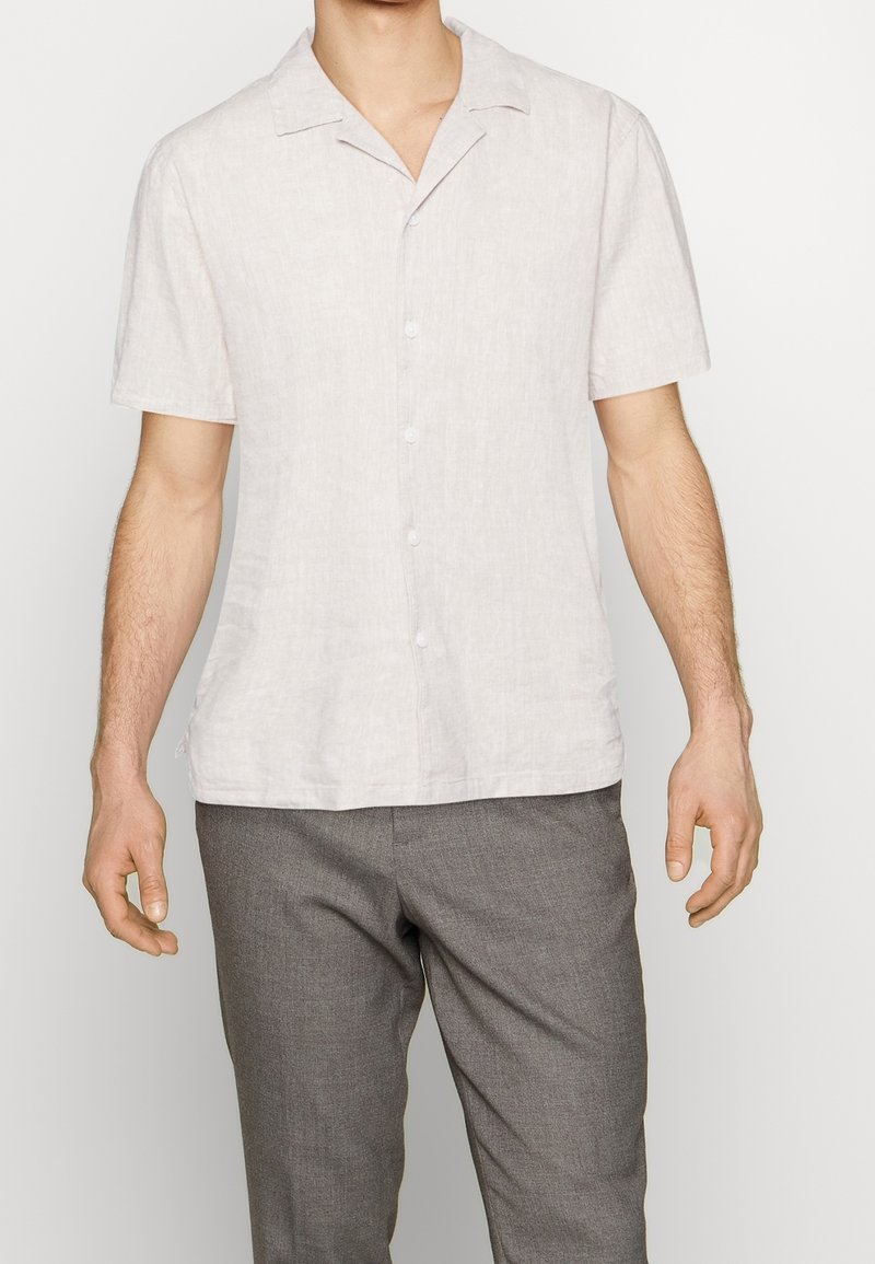 Chemise en lin de couleur claire à manches courtes, dotée d'une fermeture boutonnée à l'avant, d'un col à revers et d'une coupe décontractée. Portée avec un pantalon gris.