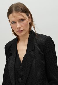 Černý blazer s texturovaným vzorem, s klopami a dvěma knoflíky. Doplněn velkými geometrickými náušnicemi, na jednobarevném pozadí.