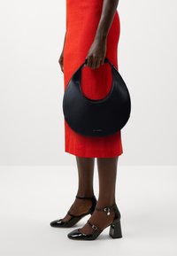 Femme en robe rouge tenant un sac à main noir en forme de croissant, portant des chaussures à talons carrés en cuir verni noir avec des brides à la cheville sur fond blanc.