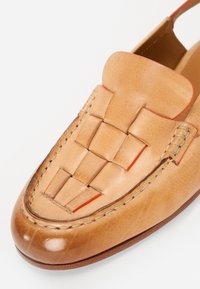 Tan skinn loafer med en vävd design på tån, kontrasterande röda detaljer och synlig sömnad längs kanterna. Slät textur.