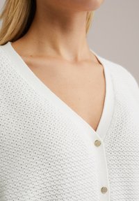 Detailní záběr osoby v texturovaném bílém pleteném svetru s zlatými knoflíky a výstřihem do V.