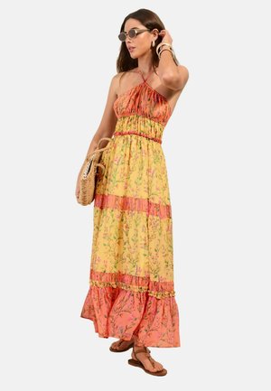 Woman wearing sleeveless, floral, tiered maxi dress in yellow and coral, carrying woven handbag, wearing sunglasses and sandals.