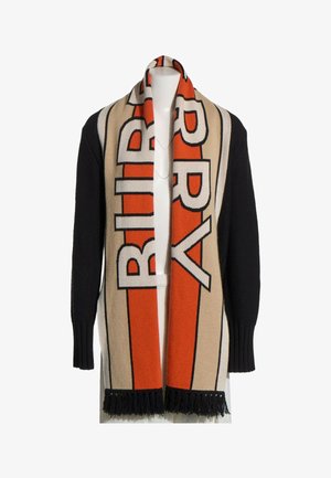 Pull noir avec une longue écharpe beige et orange ornée de grandes lettres en gras et de franges noires aux extrémités, portée sur les épaules.