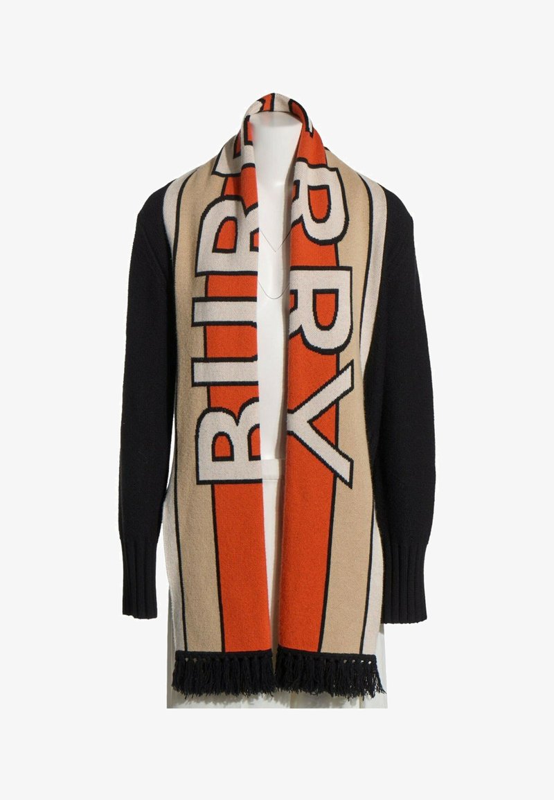 Pull noir avec une longue écharpe beige et orange ornée de grandes lettres en gras et de franges noires aux extrémités, portée sur les épaules.
