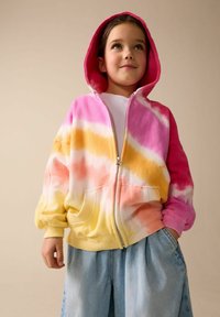 Giacca con cappuccio effetto tie-dye che sfuma dal rosa all'arancione al giallo, con zip frontale e due tasche laterali. Abbinata a pantaloncini di jeans color azzurro chiaro.