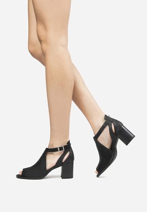 Piernas con zapatos negros de tacón grueso con puntera abierta y tiras recortadas en el tobillo sobre un fondo blanco, un pie levantado de puntillas.